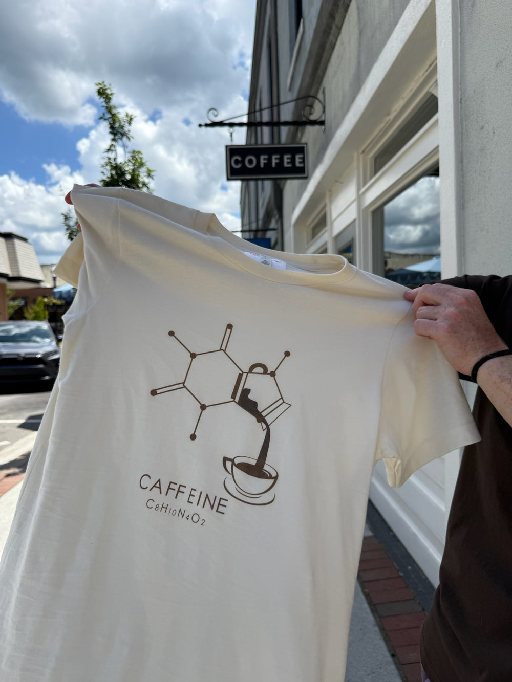 Cream Enerjava t-shirt with 'CAFFEINE' molecule graphic design held in front of a coffee shop.