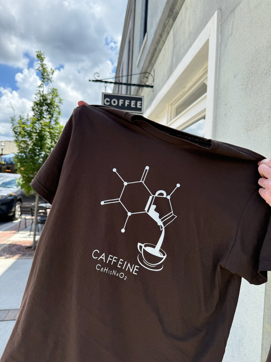 Brown Enerjava t-shirt with caffeine molecule design held in front of a coffee shop.