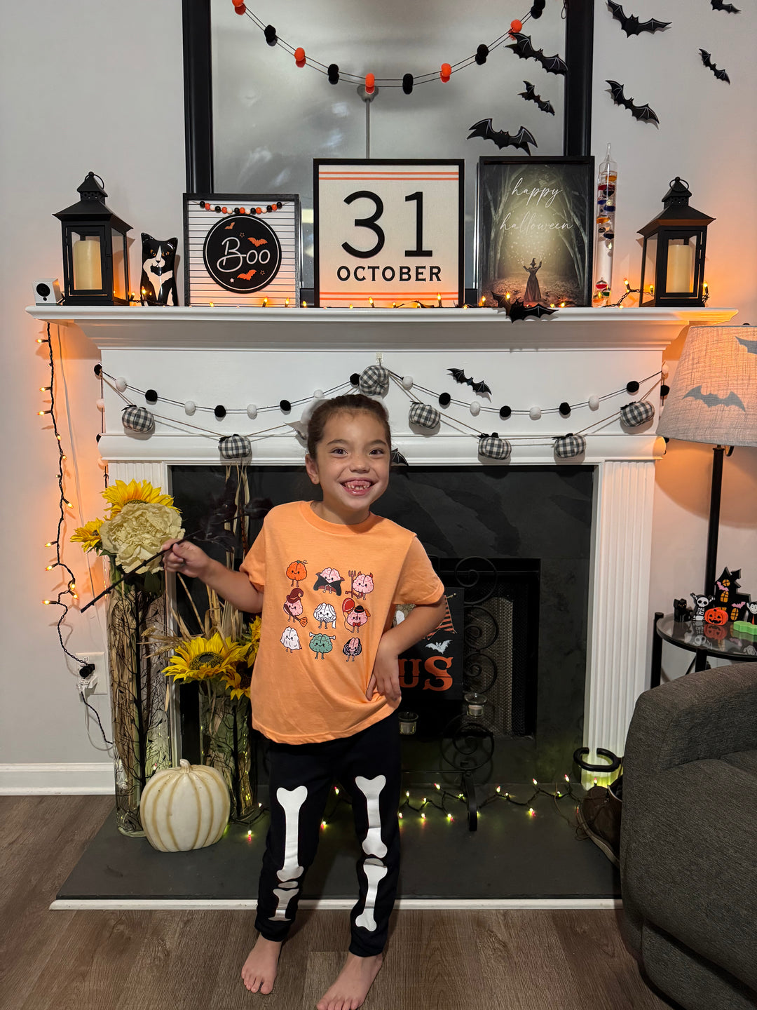 Child wearing orange Halloween Brains tee in a Halloween-themed room with decorated fireplace and 'Happy 31 October' sign.