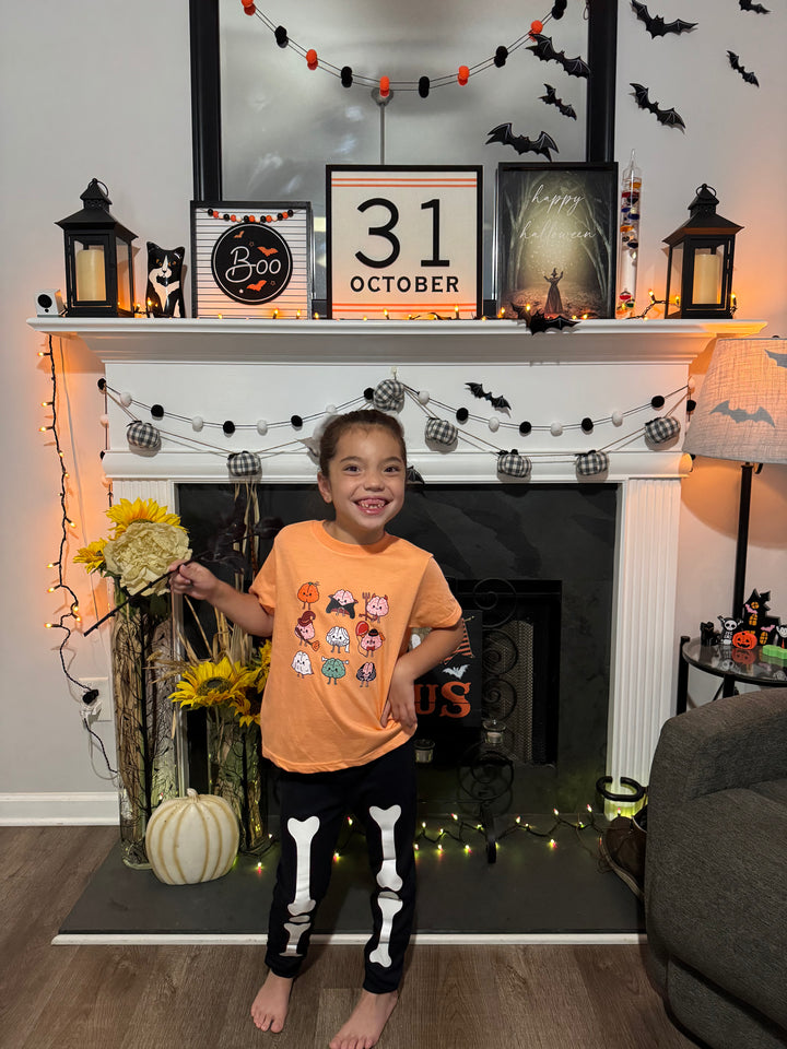Child wearing orange Halloween Brains tee in a Halloween-themed room with decorated fireplace and 'Happy 31 October' sign.