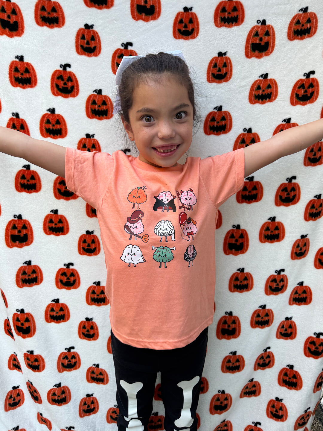 A child wearing an orange Halloween Brains shirt with cartoon brains wearing traditional Halloween costumes like a witch, a ghost, and a vampire.