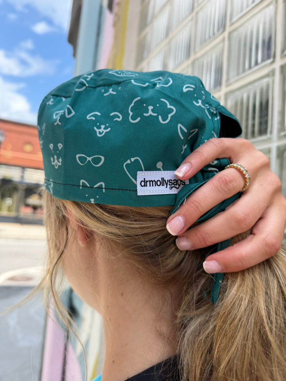 Person wearing a green scrub hat with an animal pattern, held by a hand, against an city background.