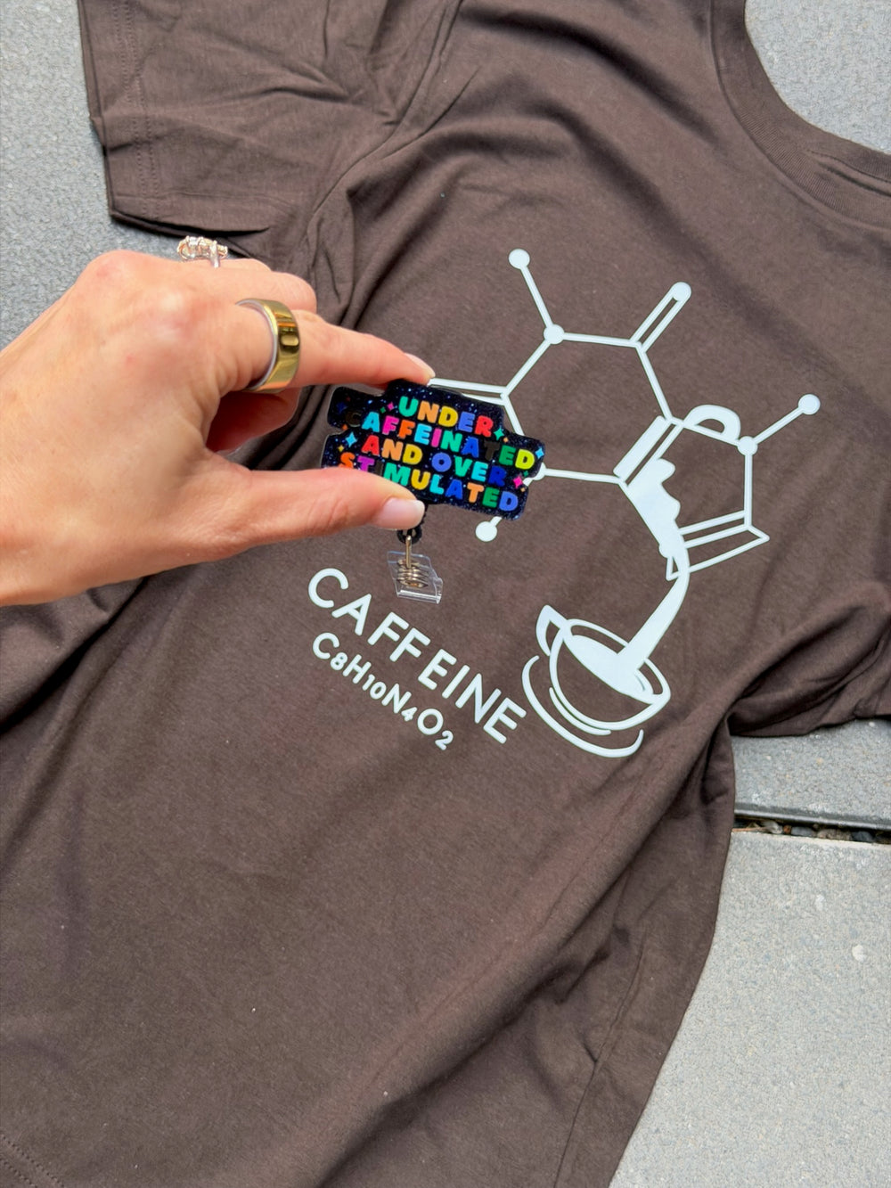 Brown t-shirt with 'CAFFEINE' molecule design and colorful badge held by a hand.