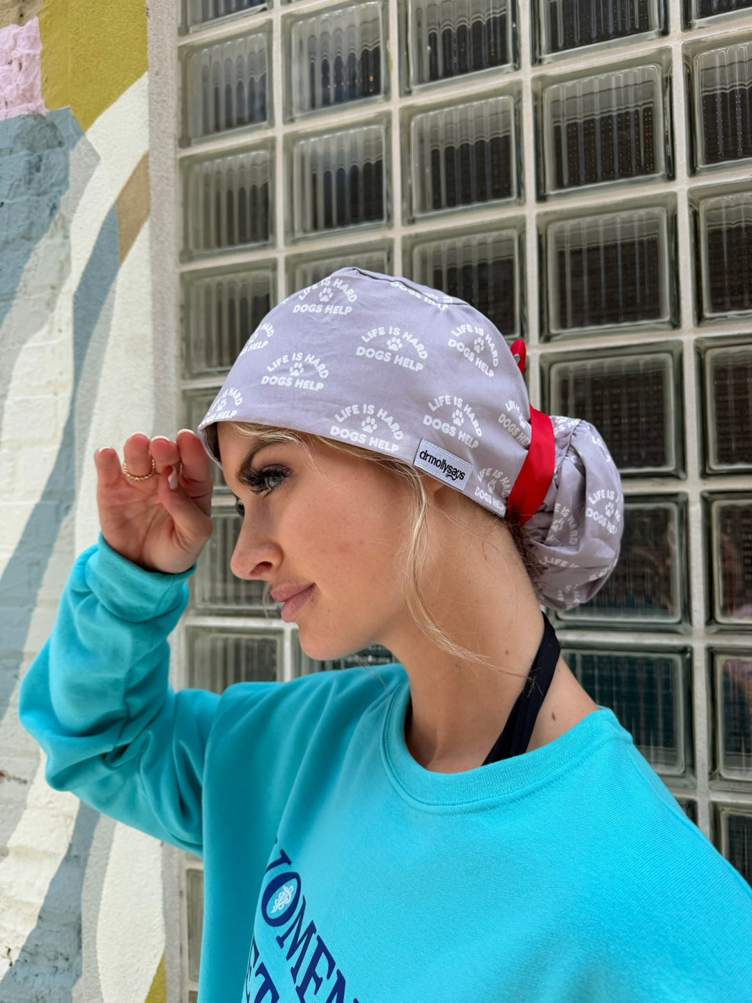Person wearing a patterned scrub hat and teal sweatshirt in front of a building. The scrub hat is gray with a red ribbon around the ponytail; the print says life is hard, dogs help.