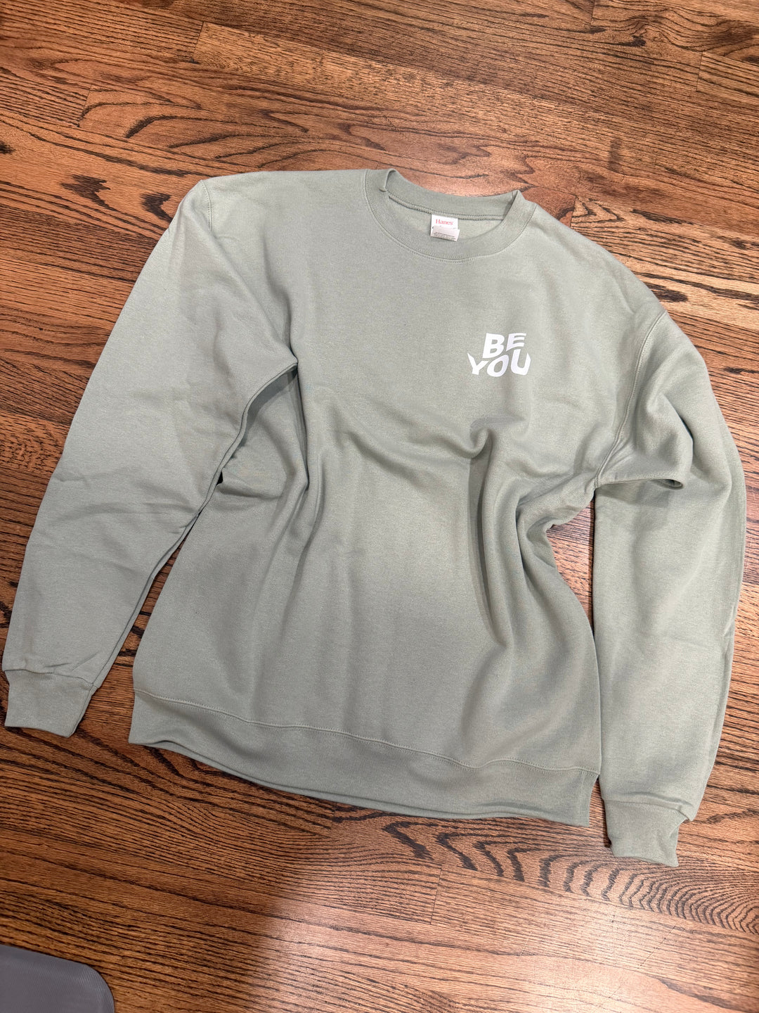 Sage green Be You sweatshirt with text on a wooden floor