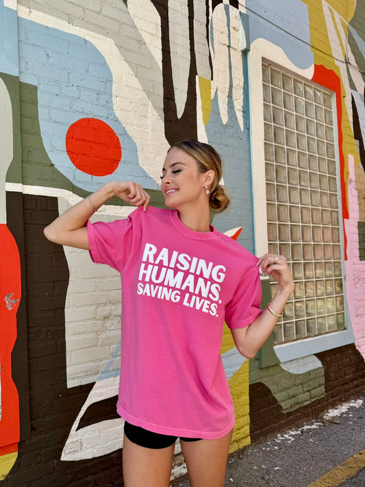 Smiling woman wearing pink Raising Humans. Saving Lives. tee by Ladyspinedoc, in front of a vibrant mural.