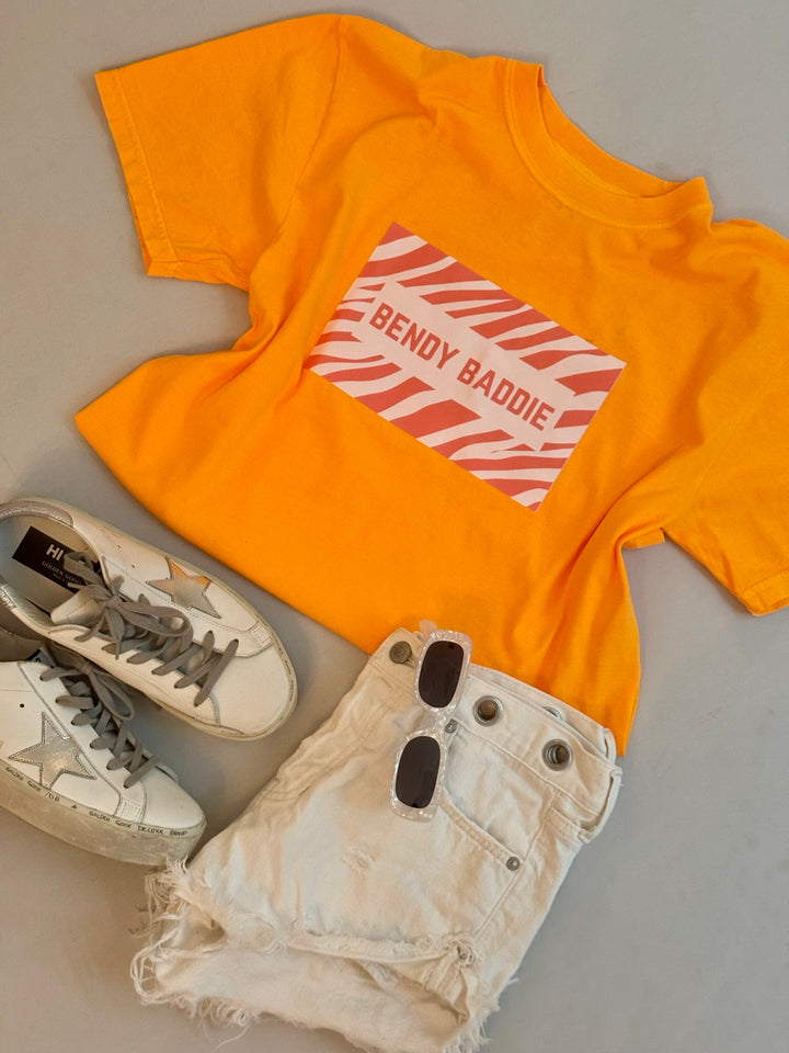 Bright orange Bendy Baddie T-shirt with pink zebra print graphic, styled with white denim shorts, black sunglasses, and white sneakers.