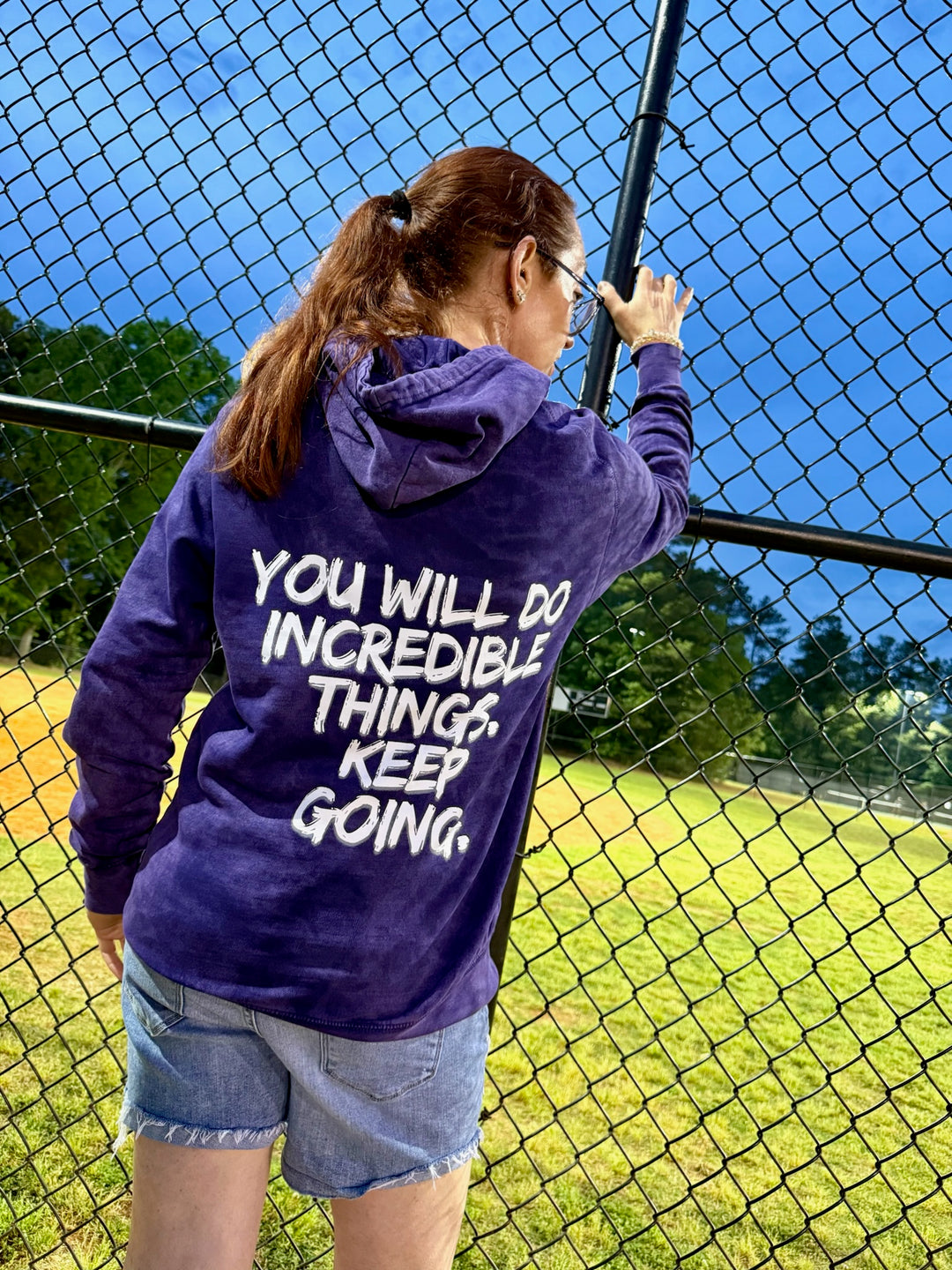 Dr. Betsy Grunch (Ladyspinedoc) wearing the purple Incredible Hoodie, displaying the back of the shirt showing bold white brushstroke message: “YOU WILL DO INCREDIBLE THINGS. KEEP GOING.”