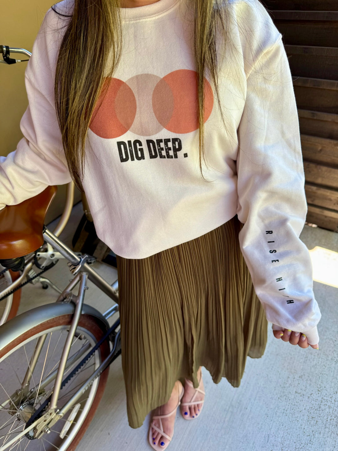 Model wearing a cream sweatshirt with 'DIG DEEP.' text on chest and "RISE HIGH" motto on sleeve and a pleated skirt, standing next to a bicycle.