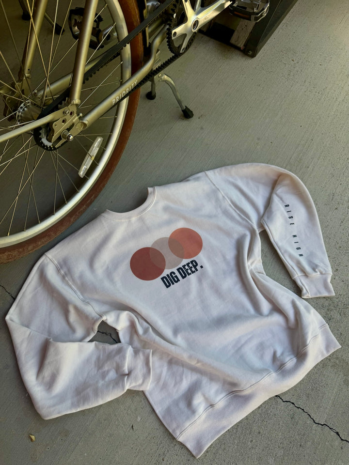 Cream long-sleeve shirt with Dig Deep graphic design on chest and Rish High sleeve text, flat laid on a concrete floor next to a bicycle.