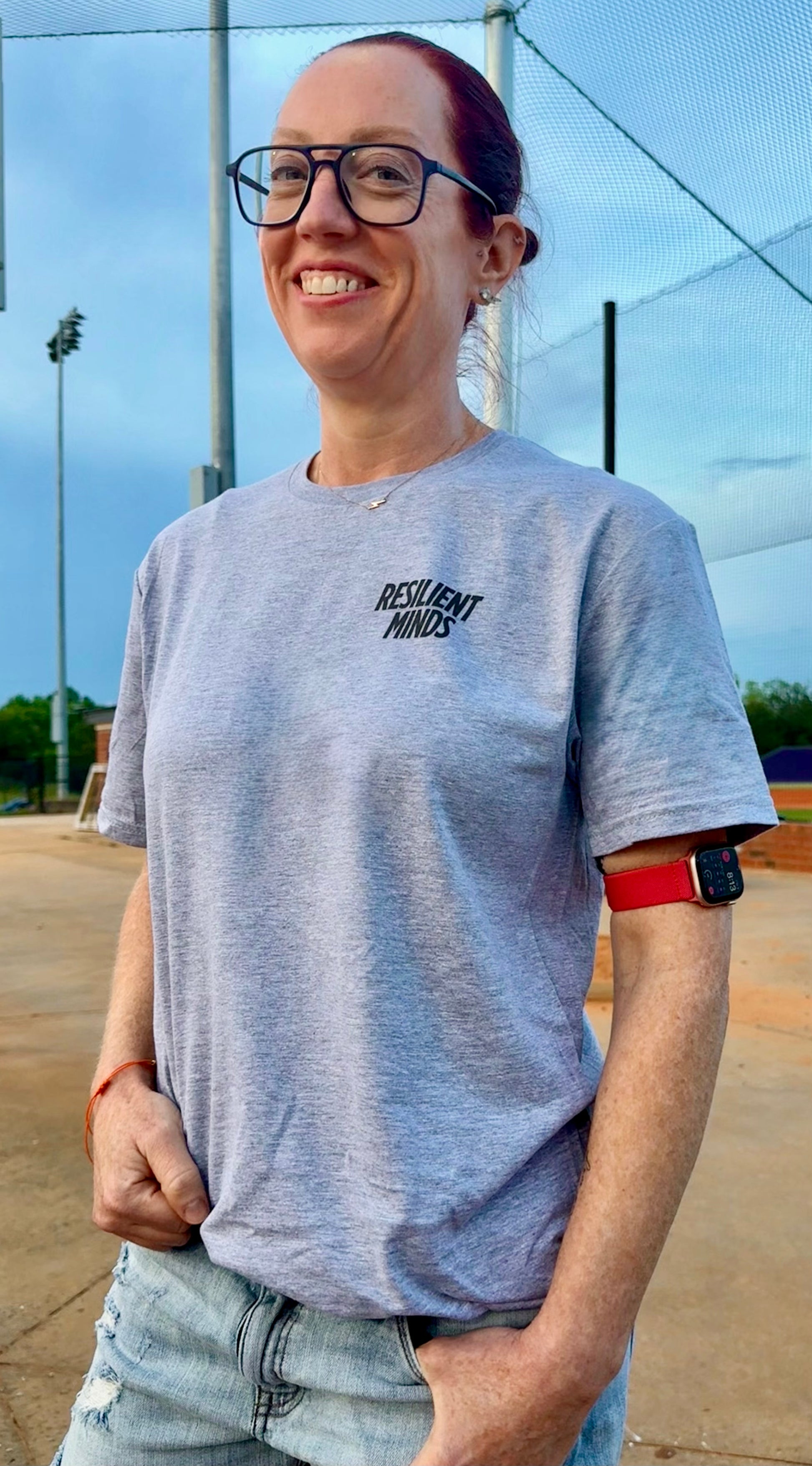 Ladyspinedoc (Dr. Betsy Grunch) wearing Gray May Brain Tumor Awareness t-shirt with “RESILIENT MINDS” printed on the left chest, standing near a baseball field.