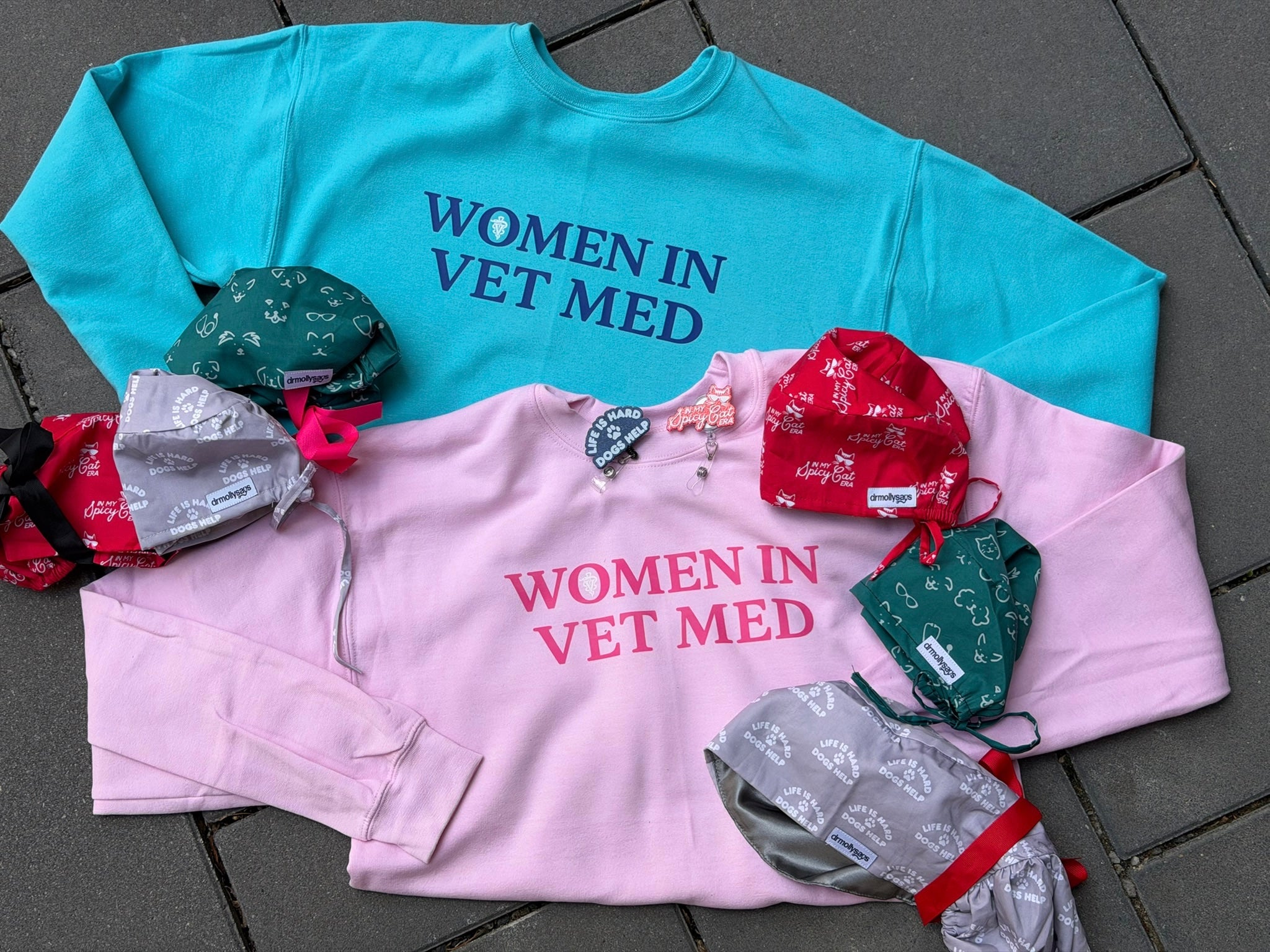 Two shirts with 'Women in Vet Med' text, surrounded by badge reels and scrub hats on a tiled floor.