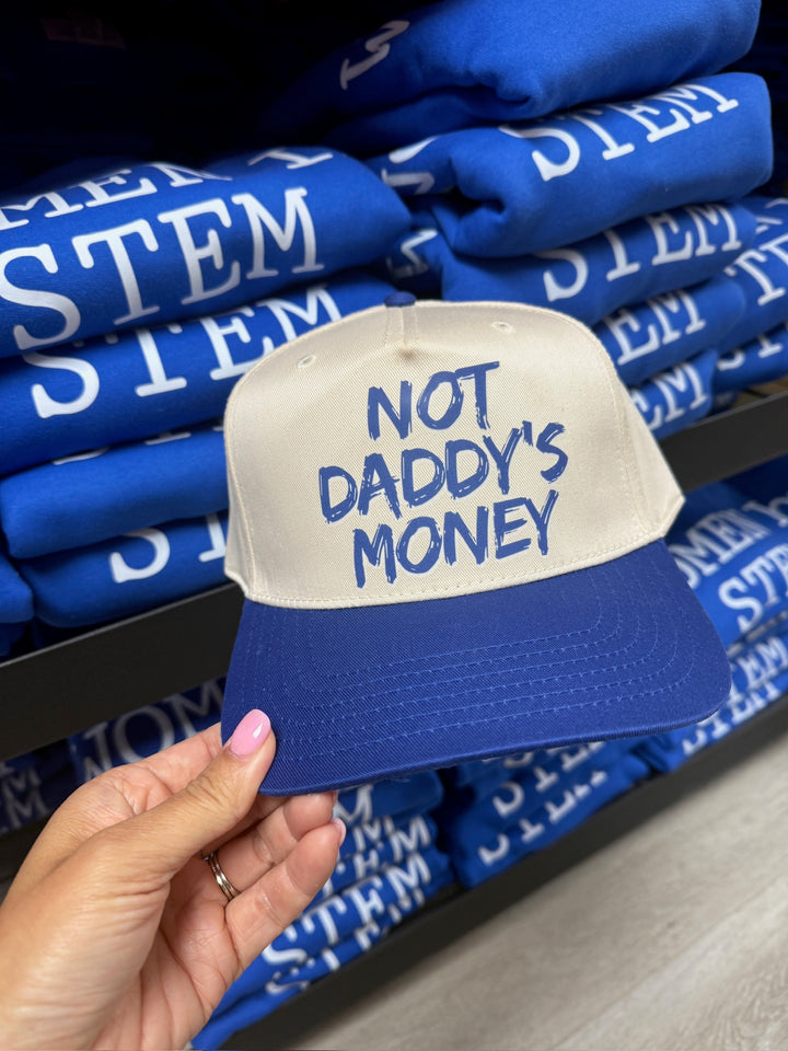 Blue and tan cap with 'Not Daddy's Money' text held in front of folded blue shirts with 'STEM' text.