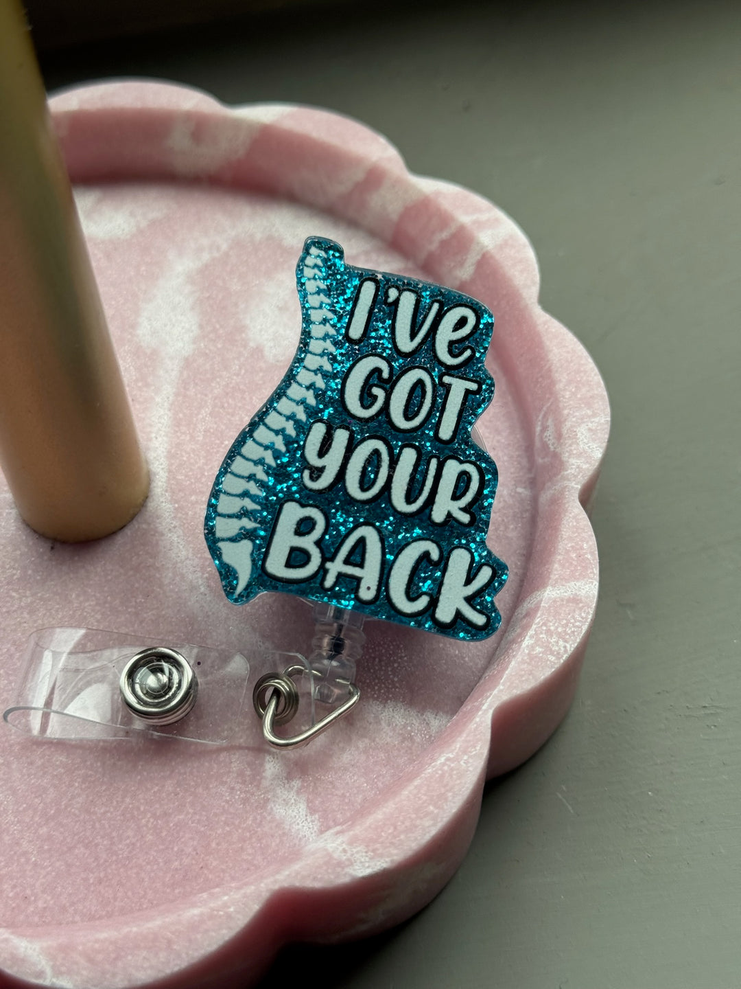 Blue glitter badge reel with spine design and motivational quote "I've Got Your Back" displayed on a pink scalloped tray.
