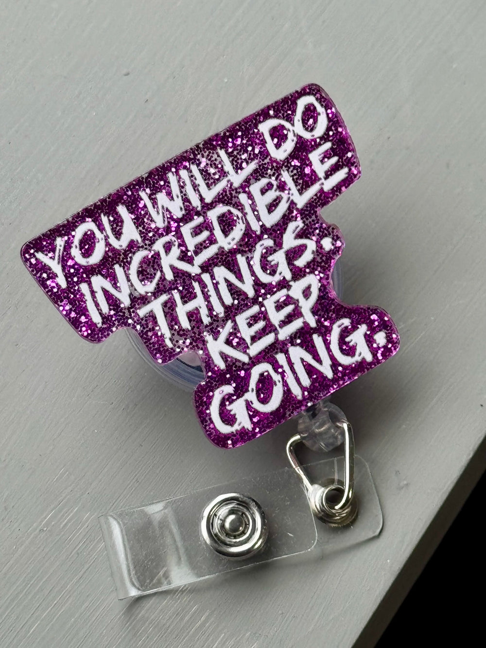 Close-up of a purple glitter badge reel reading “YOU WILL DO INCREDIBLE THINGS. KEEP GOING.” on a gray surface, highlighting its bold white lettering and retractable clip.