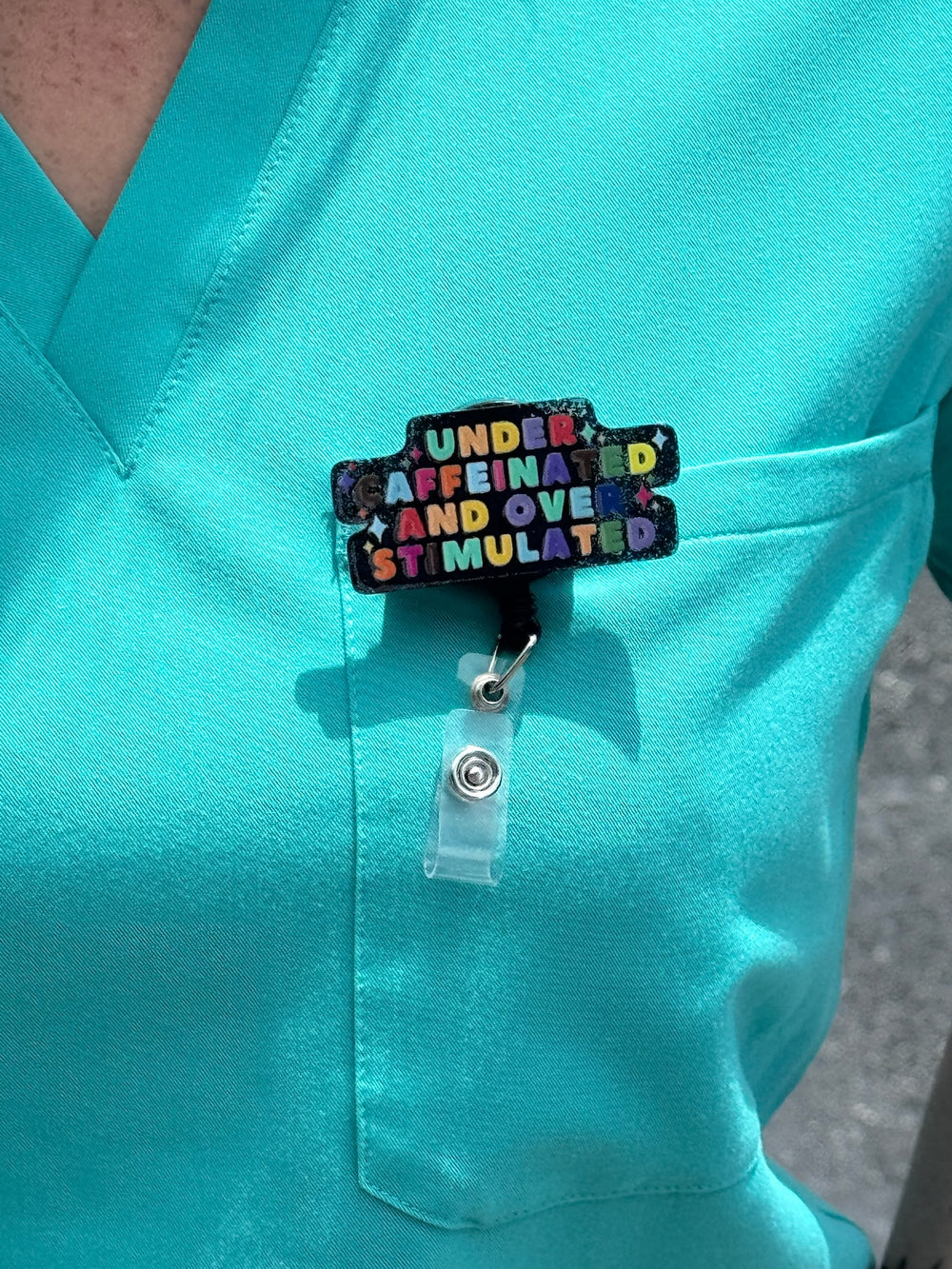 Close up of Under Caffeinated and Over Stimulated Badge Reel clipped on scrub shirt