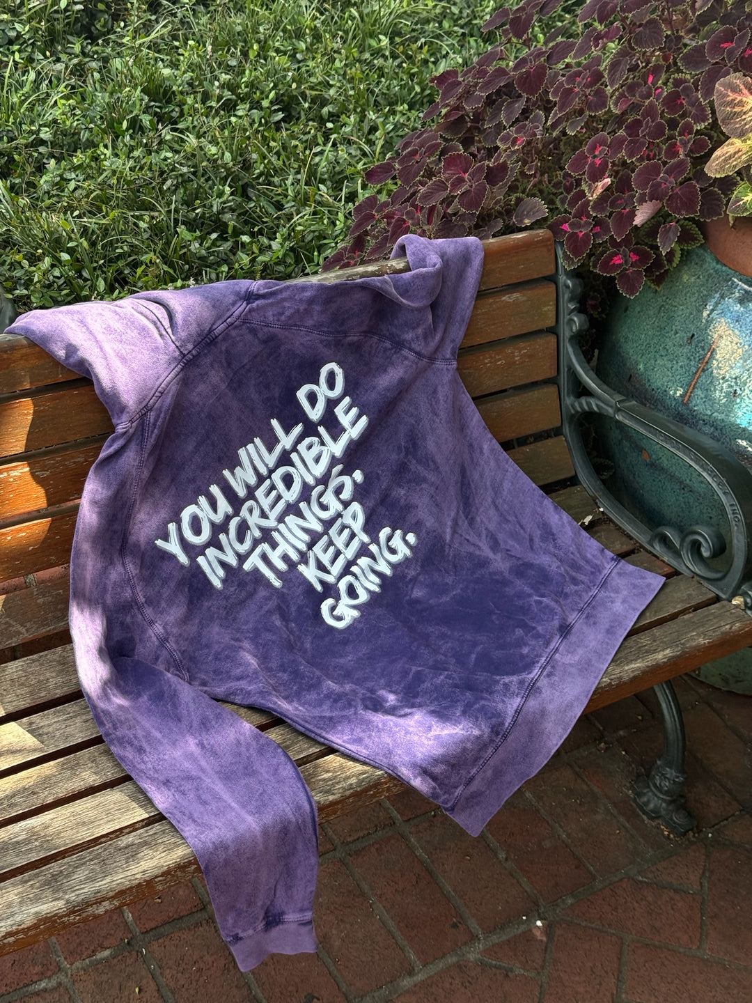 The purple Incredible Hoodie on a park bench displaying the back of the shirt showing bold white brushstroke message: “YOU WILL DO INCREDIBLE THINGS. KEEP GOING.”
