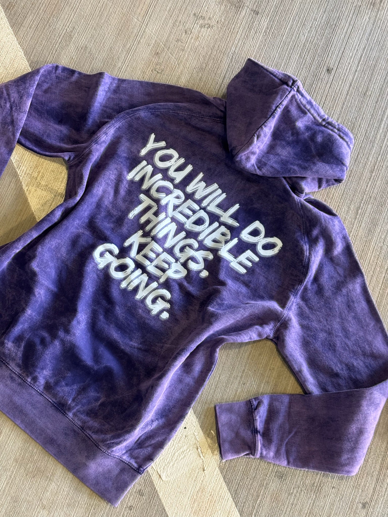 Flat lay of the purple Incredible Hoodie, displaying the back of the shirt showing bold white brushstroke message: “YOU WILL DO INCREDIBLE THINGS. KEEP GOING.”