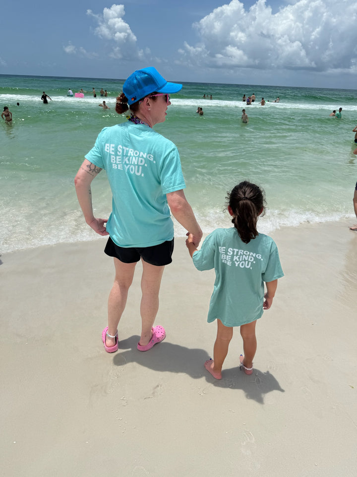 Ladyspinedoc and a young girl hold hands at the beach, both wearing matching aqua blue T-shirts with the "BE STRONG. BE KIND. BE YOU." message on the back, gazing out at the waves together.