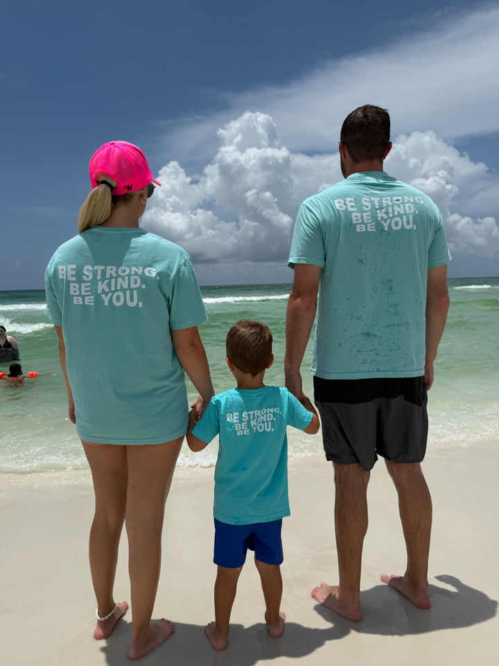 A family of three—mom, dad, and young son—stands hand-in-hand at the shoreline, all wearing matching aqua blue "BE STRONG. BE KIND. BE YOU." T-shirts, looking out at the ocean under a partly cloudy sky.