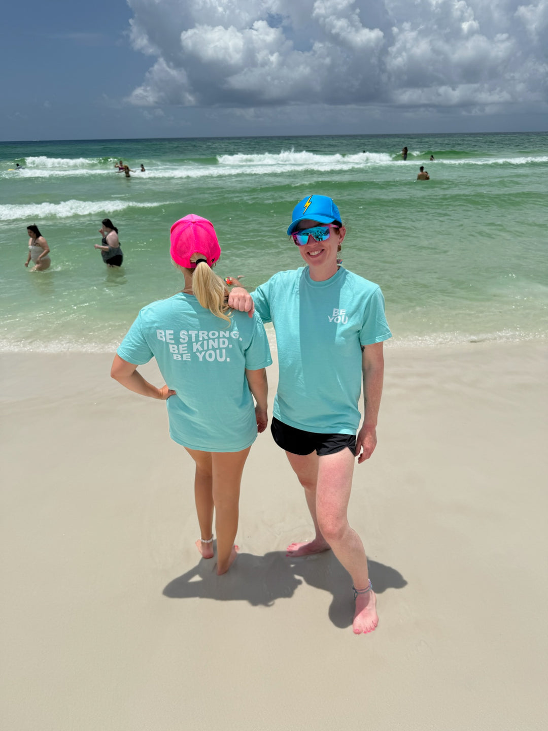 Ladyspinedoc and friend stand on the beach, one facing the camera and smiling while the other shows off the back of her shirt, both wearing matching "BE YOU" T-shirts in aqua blue.