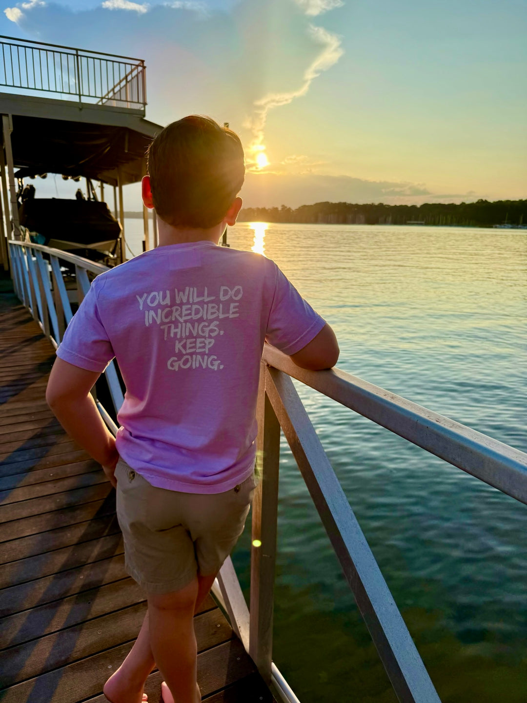 Back of kid facing sunset at a lake wearing the violet Incredible Youth Tee with the motivational words "You will do incredible things. Keep Going" displayed on the back. 