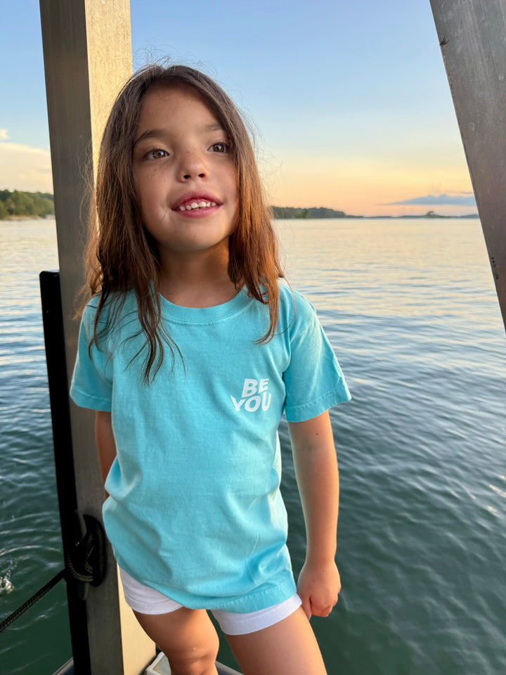 Close-up of a young girl in the Be You Youth Tee by Ladyspinedoc, standing by the water with the front logo “BE YOU” in white.