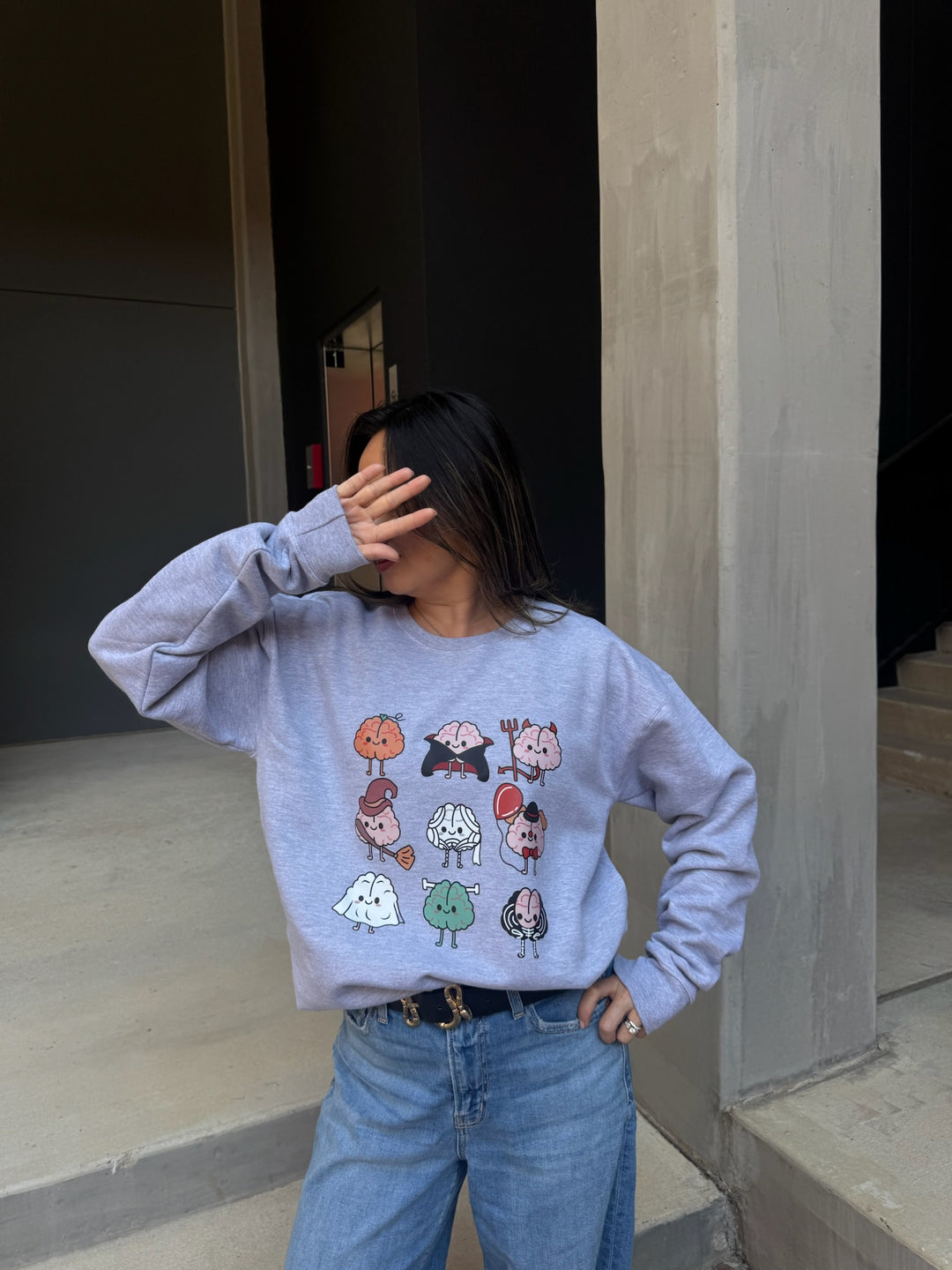 Model with hand on face wearing Gray Halloween Brains Crewneck Sweatshirt with Cartoon Brain Characters: Pumpkin, Ghost, Witch, Vampire, Mummy & More 