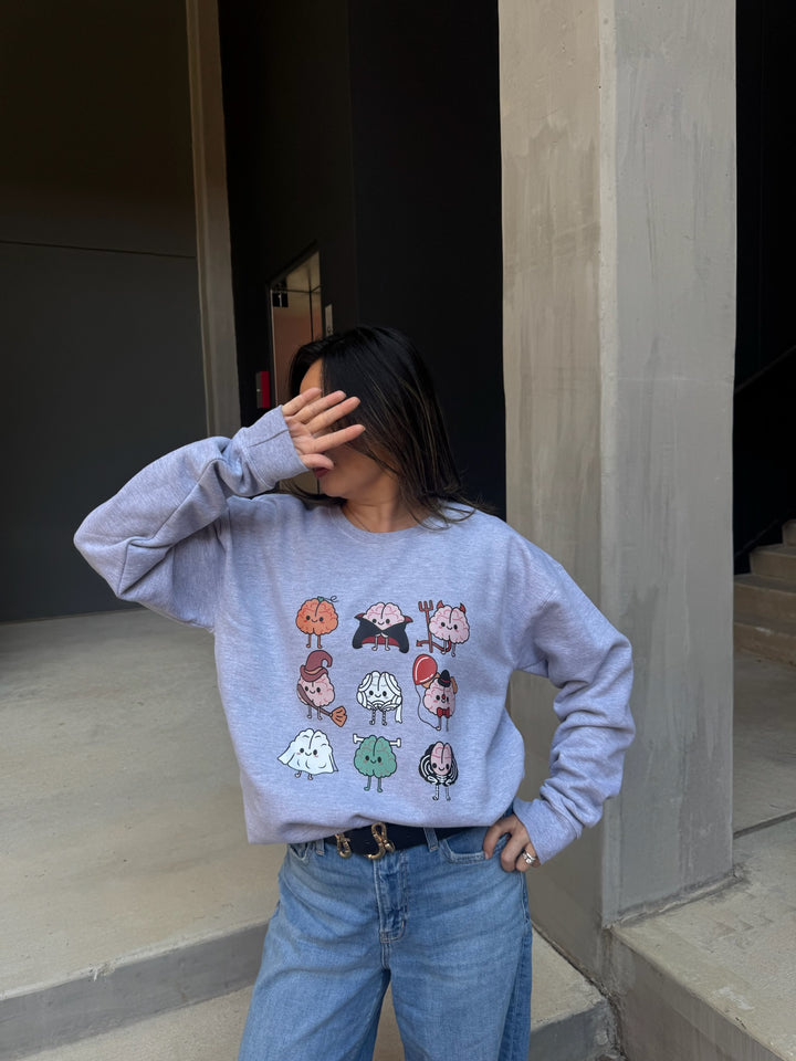 Model with hand on face wearing Gray Halloween Brains Crewneck Sweatshirt with Cartoon Brain Characters: Pumpkin, Ghost, Witch, Vampire, Mummy & More 