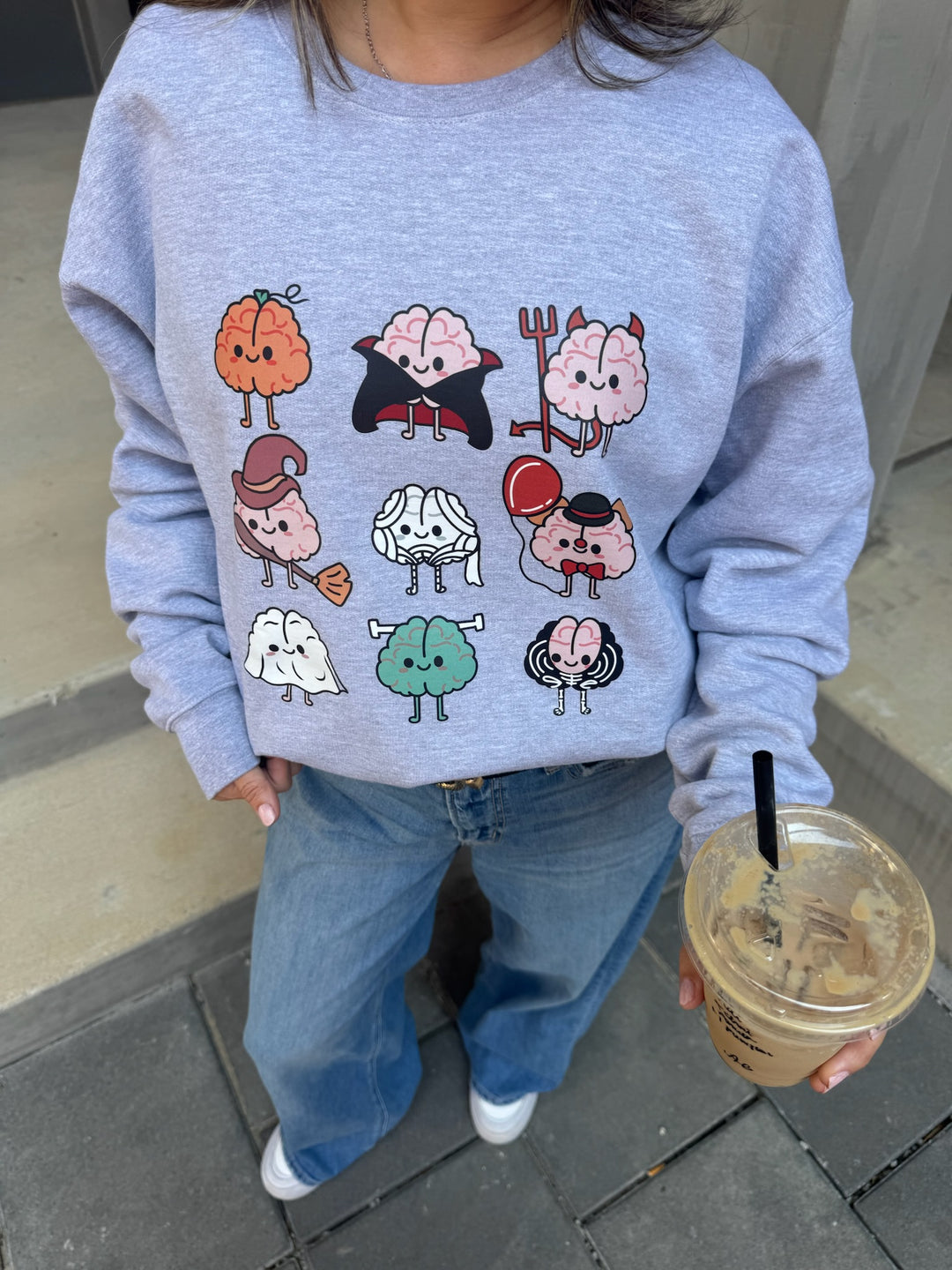 Model wearing Gray Halloween Brains Crewneck Sweatshirt with Cartoon Brain Characters: Pumpkin, Ghost, Witch, Vampire, Mummy & More and holding an iced coffee