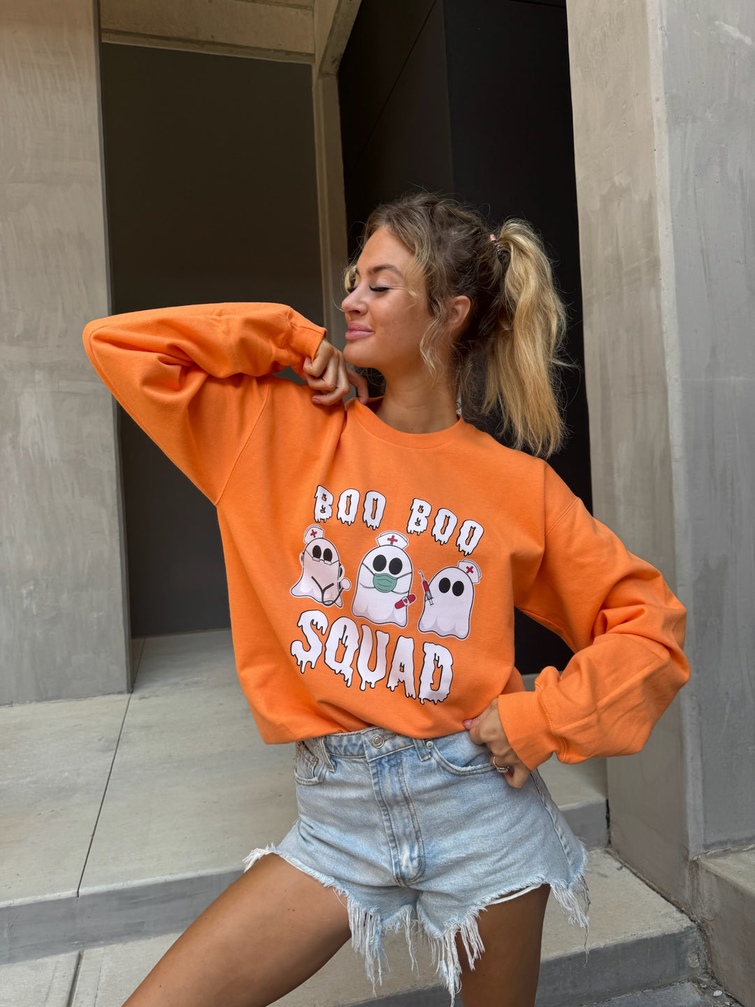 Smiling model wearing cut offs and Ladyspinedoc x Dr. Beachgem Collab: Boo Boo Squad Crewneck Sweatshirt – Halloween Shirt for Healthcare Heroes with ghost nurses