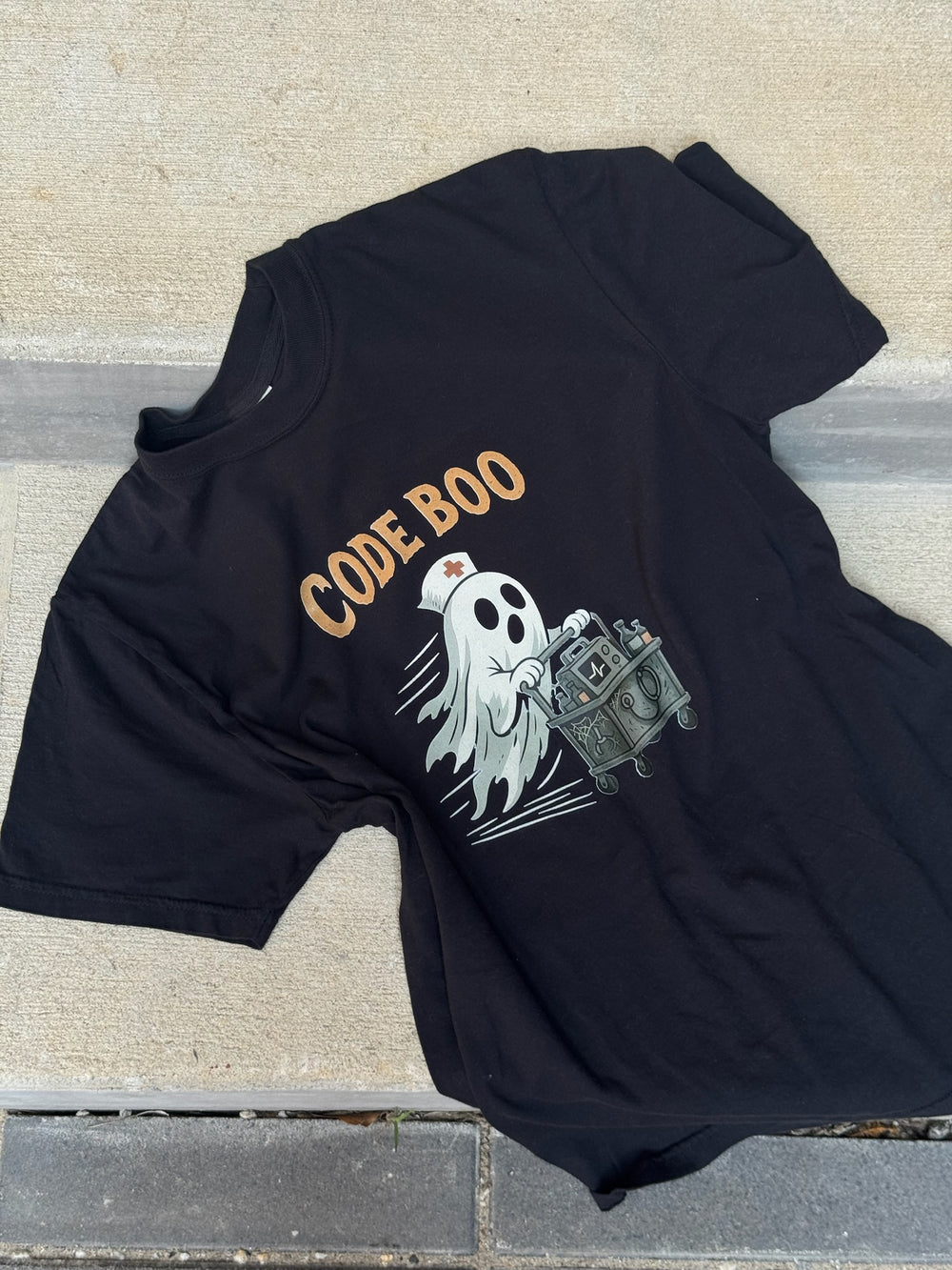 Flat lay of the black Scrubhacks Collab: Code Boo Tee – Halloween Shirt for Healthcare Heroes. It shows a nurse ghost with a creepy code cart.