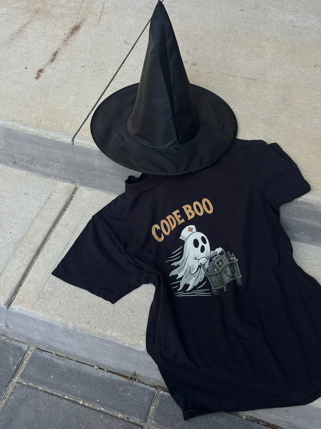 Black Scrubhacks Collab: Code Boo Tee  next to a black witch hat. Shirt design shows a nurse ghost with a creepy code cart.