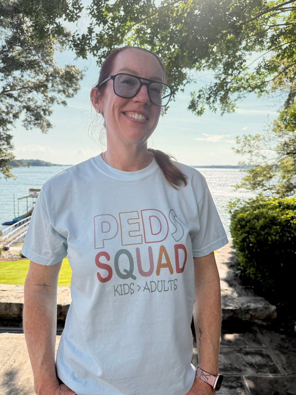 Close-up of Dr. Betsy Grunch smiling in her “PEDS SQUAD – KIDS > ADULTS” shirt. The lake sparkles in the background as sunlight filters through the trees overhead.