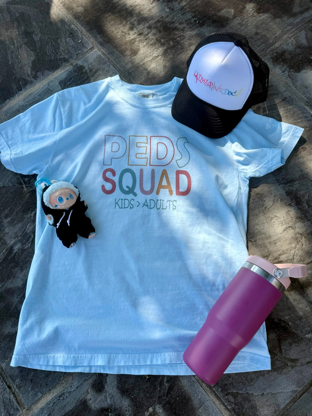 Flat lay of the pastel blue “PEDS SQUAD – KIDS > ADULTS” t-shirt on a stone surface, alongside a Ladyspinedoc logo trucker hat, a small plush doll, and a pink water tumbler.