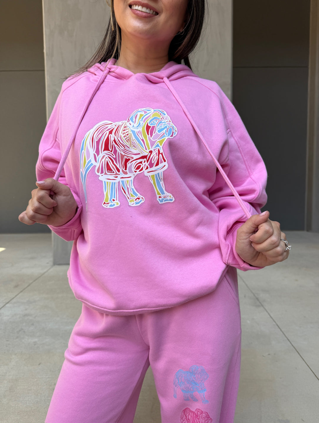 Model wearing Chan Art Collab: Game Day Set - Pink Hoodie & Sweatpants while pulling on the drawstrings of the hoody. Design shows colorful bulldog on front of shirt.