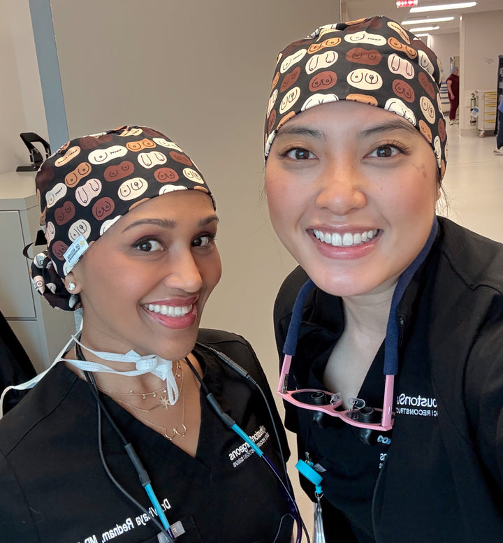 Two smiling female surgeons wearing matching Bosom Cap scrub hats featuring illustrated boobs in various shapes and skin tones, inside a hospital setting.