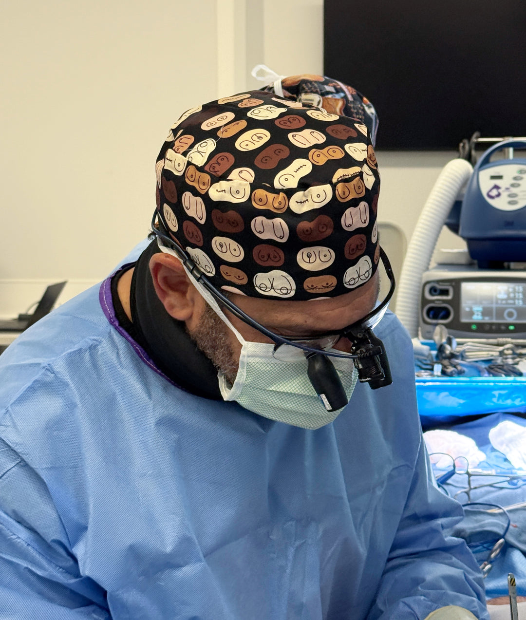 Surgeon wearing The Bosom Cap scrub hat featuring a diverse boob print in shades of brown, tan, and cream, while performing surgery in an operating room.